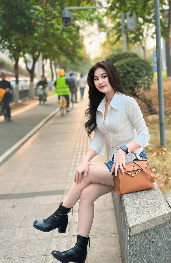 A woman sits on a stone ledge on a city sidewalk, wearing a white long-sleeve button-up shirt, denim shorts, black ankle boots, and holding a brown handbag.