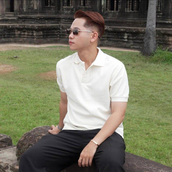 A man wearing a white short-sleeve polo shirt and black pants sits outdoors on a stone bench in front of a grassy lawn and historic stone building.