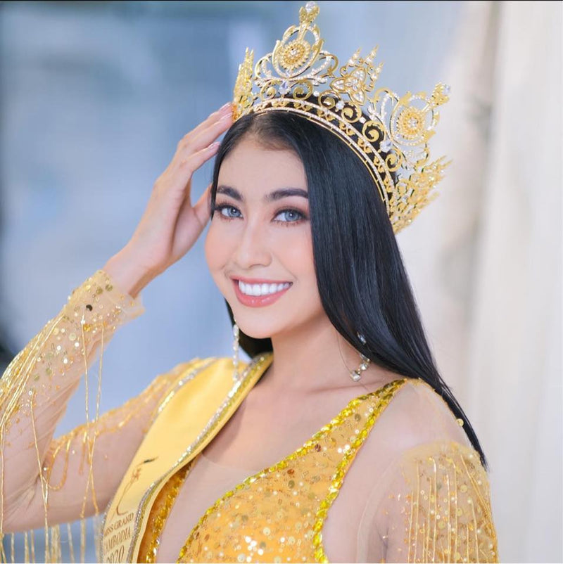 Woman in a gold outfit with a crown, smiling against a blurred background