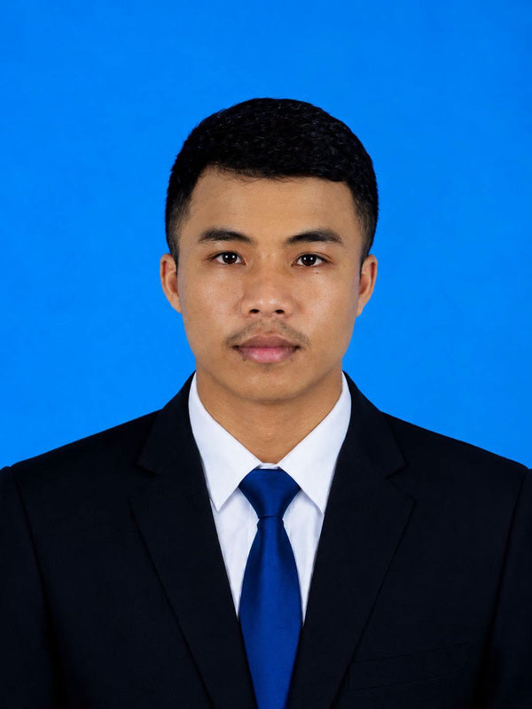 A man wearing a dark navy suit jacket, white dress shirt, and blue tie, photographed against a solid blue background.