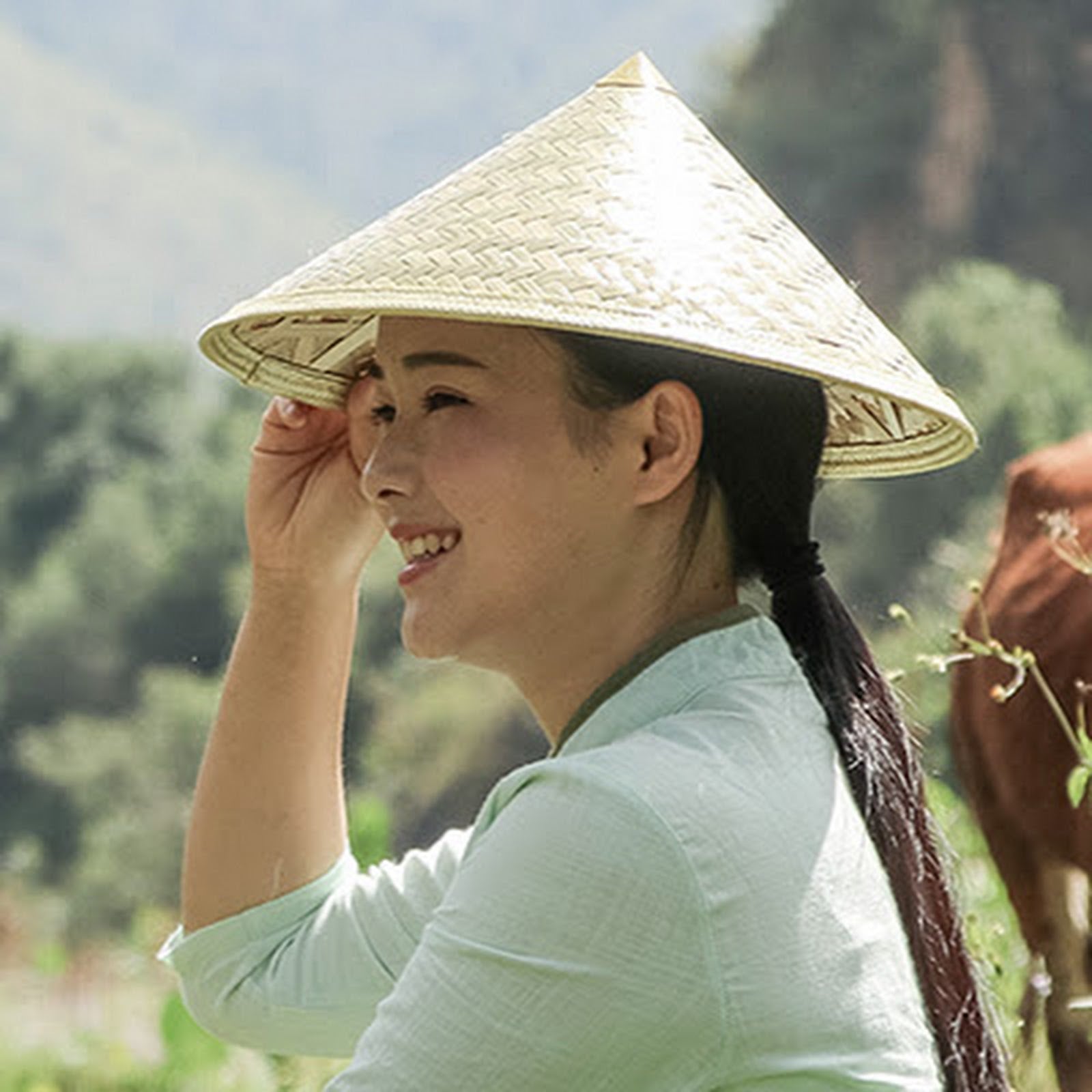 A woman wearing a traditional woven conical straw hat and a light mint green top, smiling outdoors with a mountain and greenery in the background.
