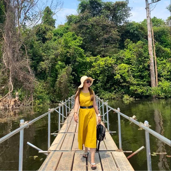 Woman in a yellow dress and hat standing on a wooden bridge over water with greenery in the background