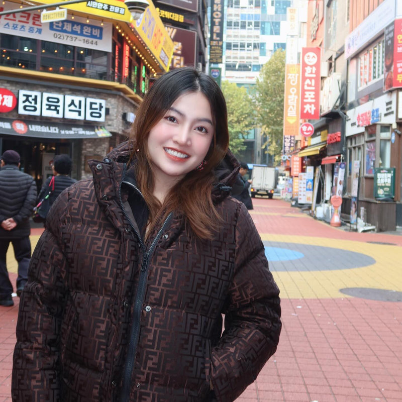 Woman in a brown patterned jacket standing on a street with shops and signs in the background