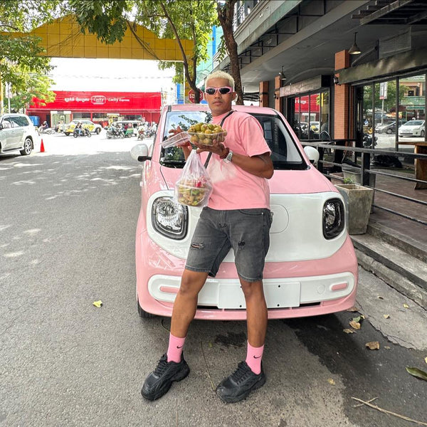 Person holding food items in front of a pink and white car on a street.