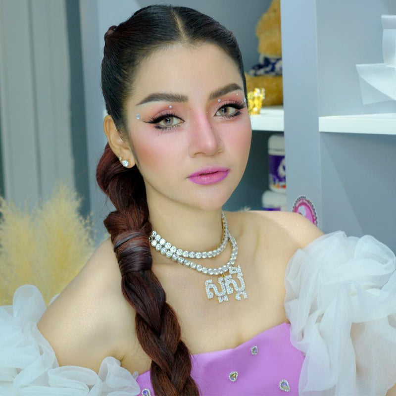 Woman with braided hair and makeup, wearing a necklace, in a blurred indoor setting.