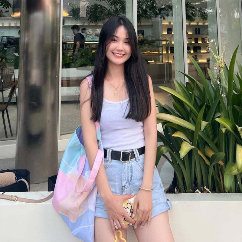 Woman in a light purple top and denim shorts holding a colorful bag, standing in an outdoor setting with plants and a building in the background.