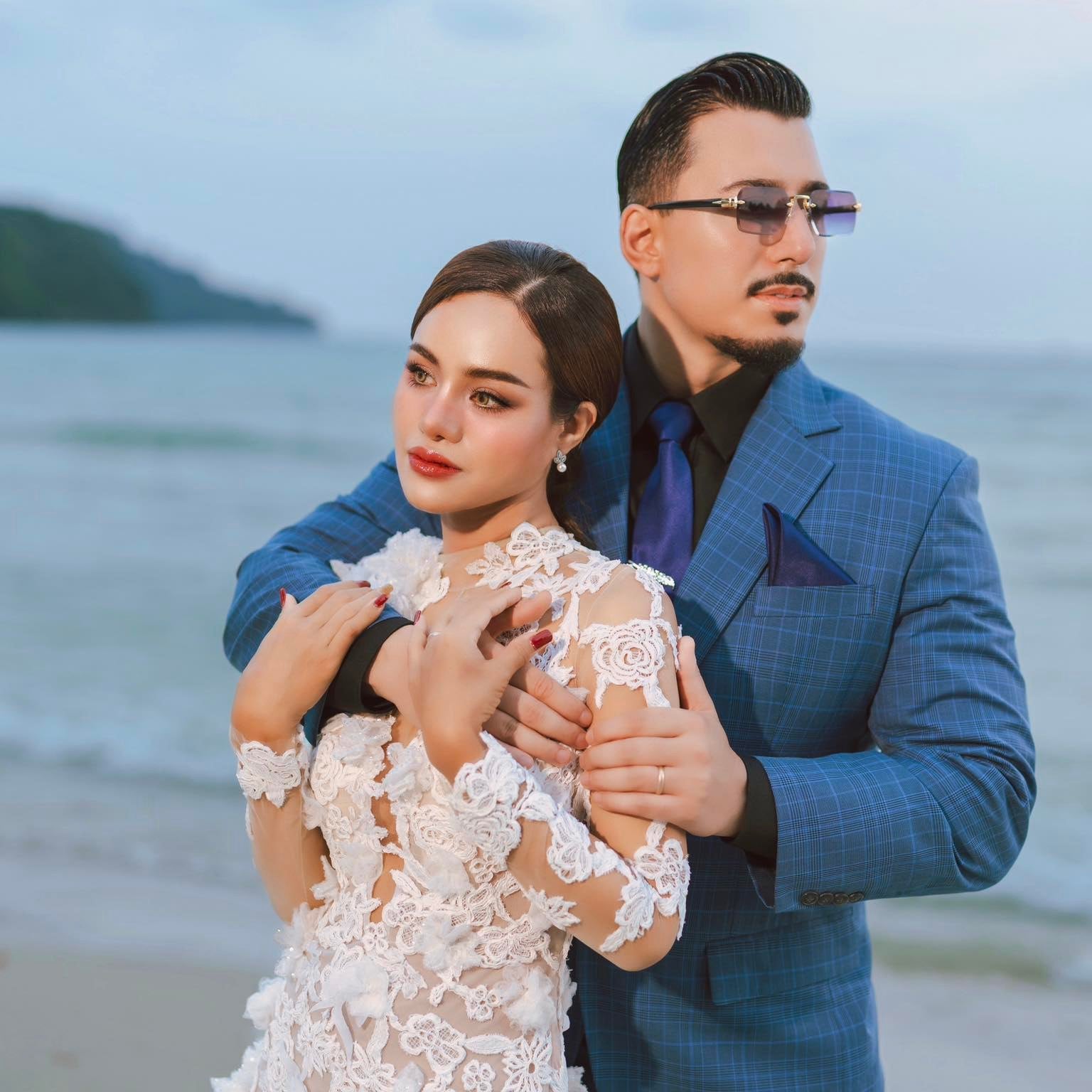 Man in a blue suit and woman in a white lace dress standing on a beach.