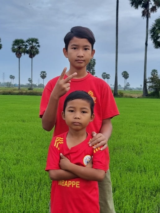 Two children in red shirts standing in a green field with palm trees in the background