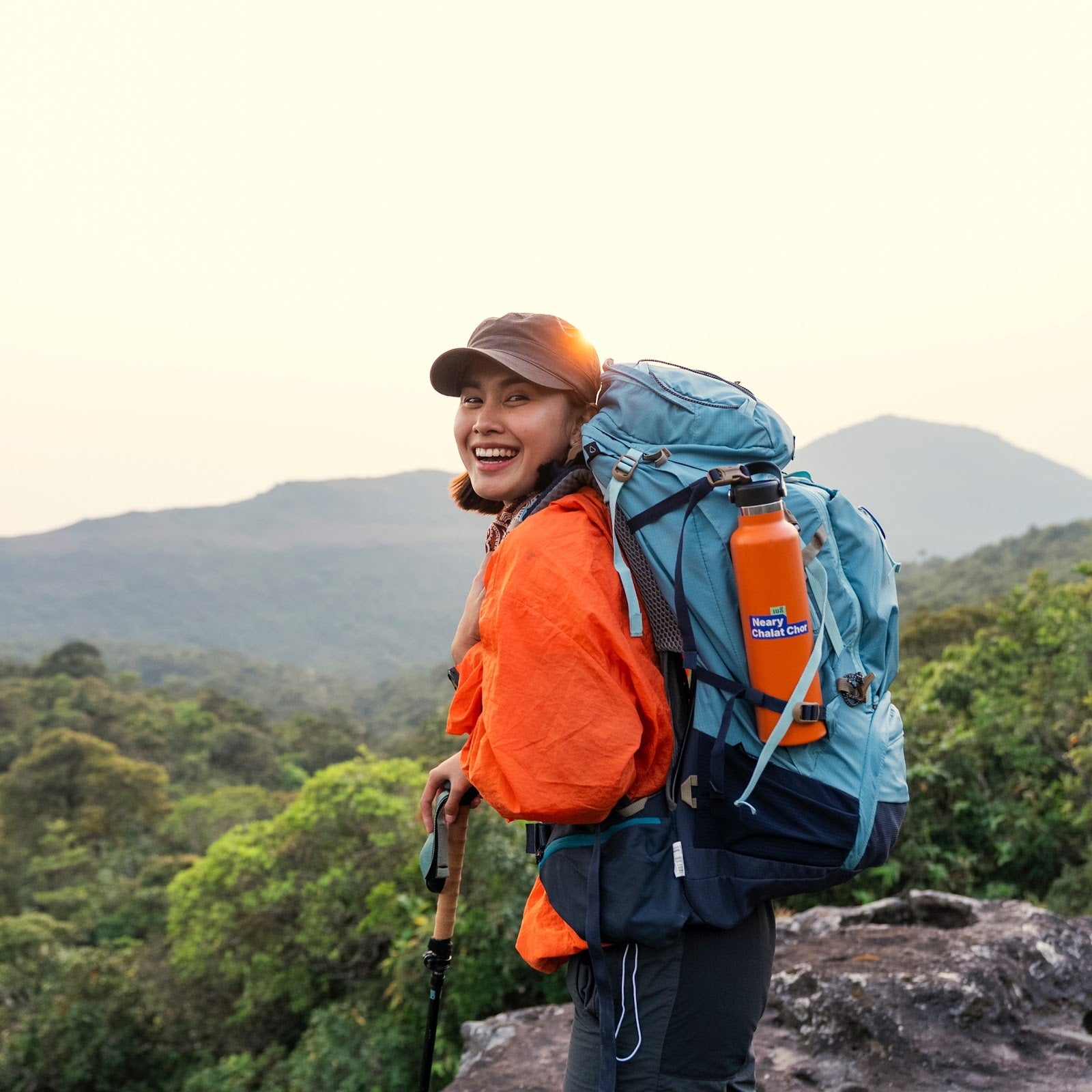 A smiling woman hiking outdoors on a mountain with a large blue backpack, orange jacket, and an orange water bottle. The background shows green forest and distant hills under a pale sky.