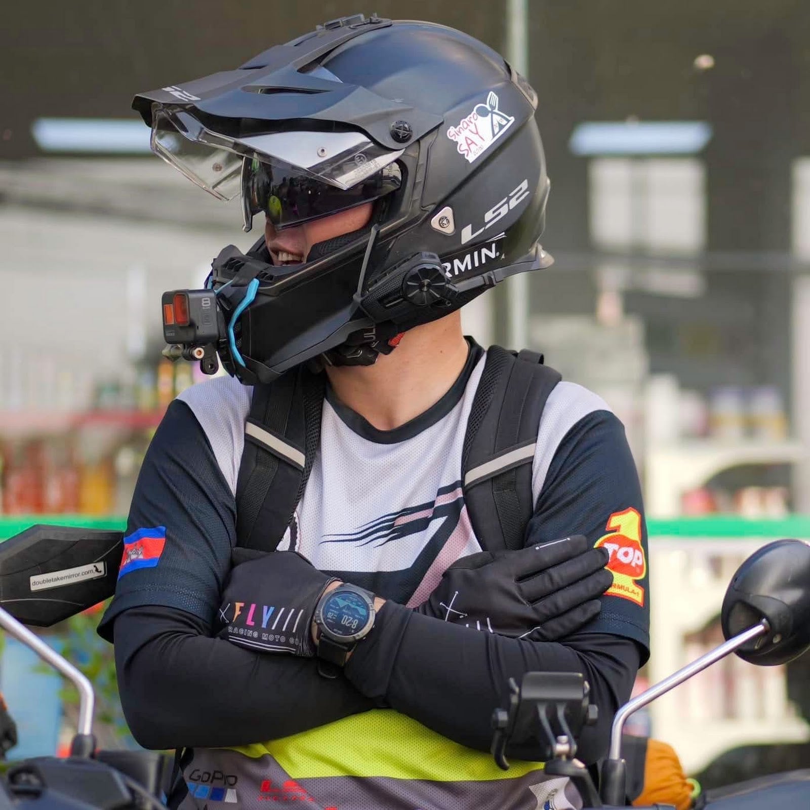 A person wearing a black full-face motorcycle helmet with a mounted camera and visor, along with a white and black long-sleeve jersey, black gloves, and a backpack, seated on a motorcycle in an outdoor setting.