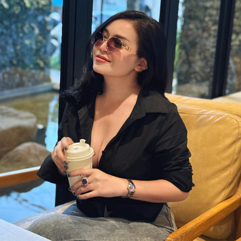 Woman sitting on a yellow couch holding a coffee cup, wearing sunglasses and a black shirt.