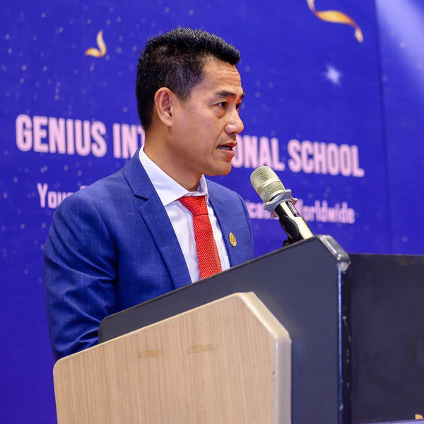 Man in a blue suit and red tie speaking at a podium with 'GENIUS INTERNATIONAL SCHOOL' in the background.