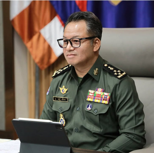 Man in military uniform with various decorations sitting at a desk with a laptop, in front of an American flag.