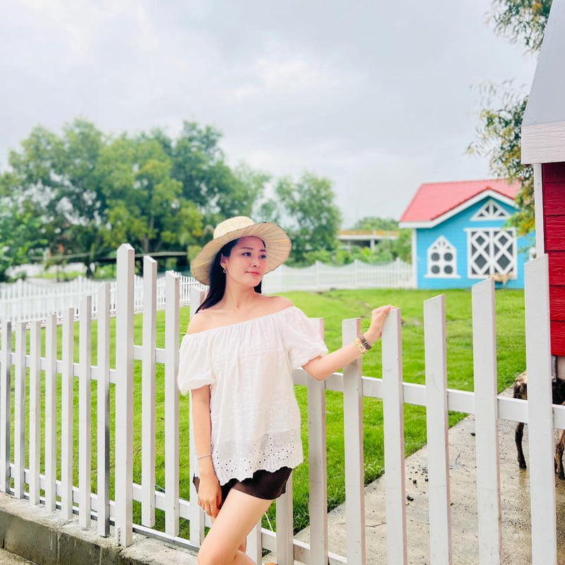 Woman in a white off-shoulder top and straw hat standing in front of a white picket fence with a colorful playhouse in the background.