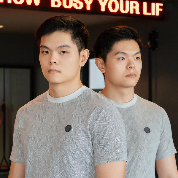 Two identical twins wearing matching gray shirts with a logo in front of a neon sign.