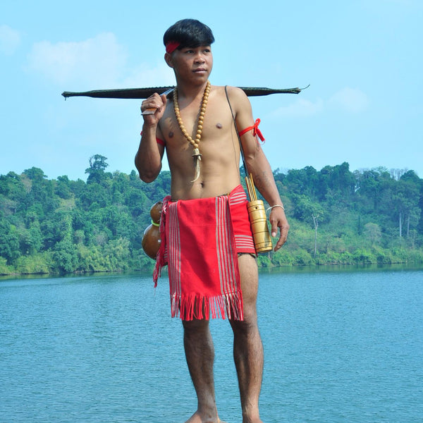 Man in traditional attire holding a bow and arrow by a body of water with trees in the background